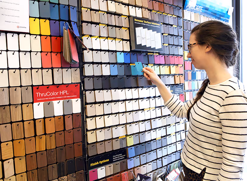 A woman with glasses and a striped shirt selects a blue color sample from a wall display of various wood and color finish samples.