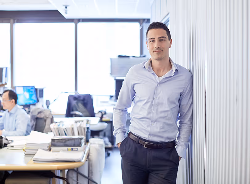 Confident man in a light blue shirt leaning against a wall in a bright office setting.