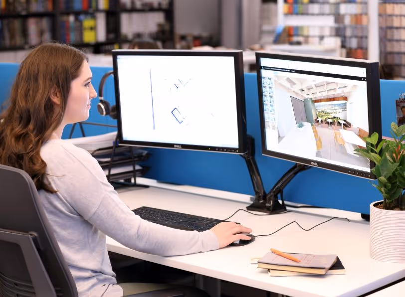Woman sitting at a desk using a dual-monitor setup displaying architectural plans and a 3D interior design rendering.