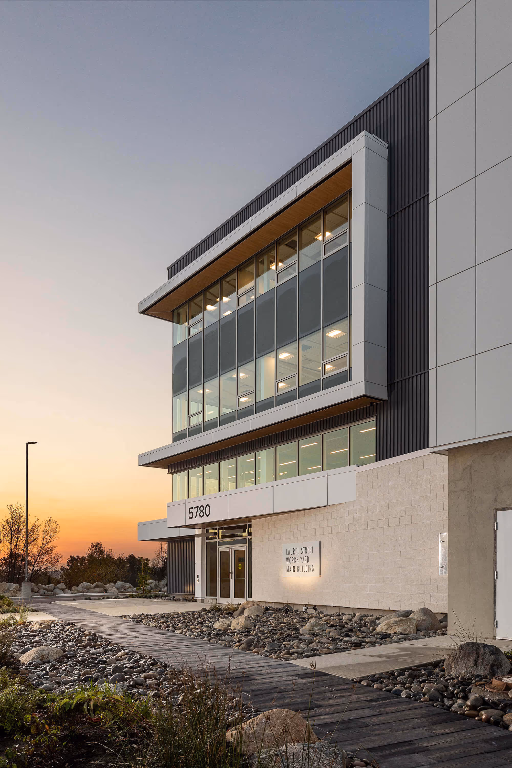 Modern office building at sunset with glass windows, a sign reading 'Laurel Street Works Yard Main Building,' and stone landscaping by a wooden pathway.