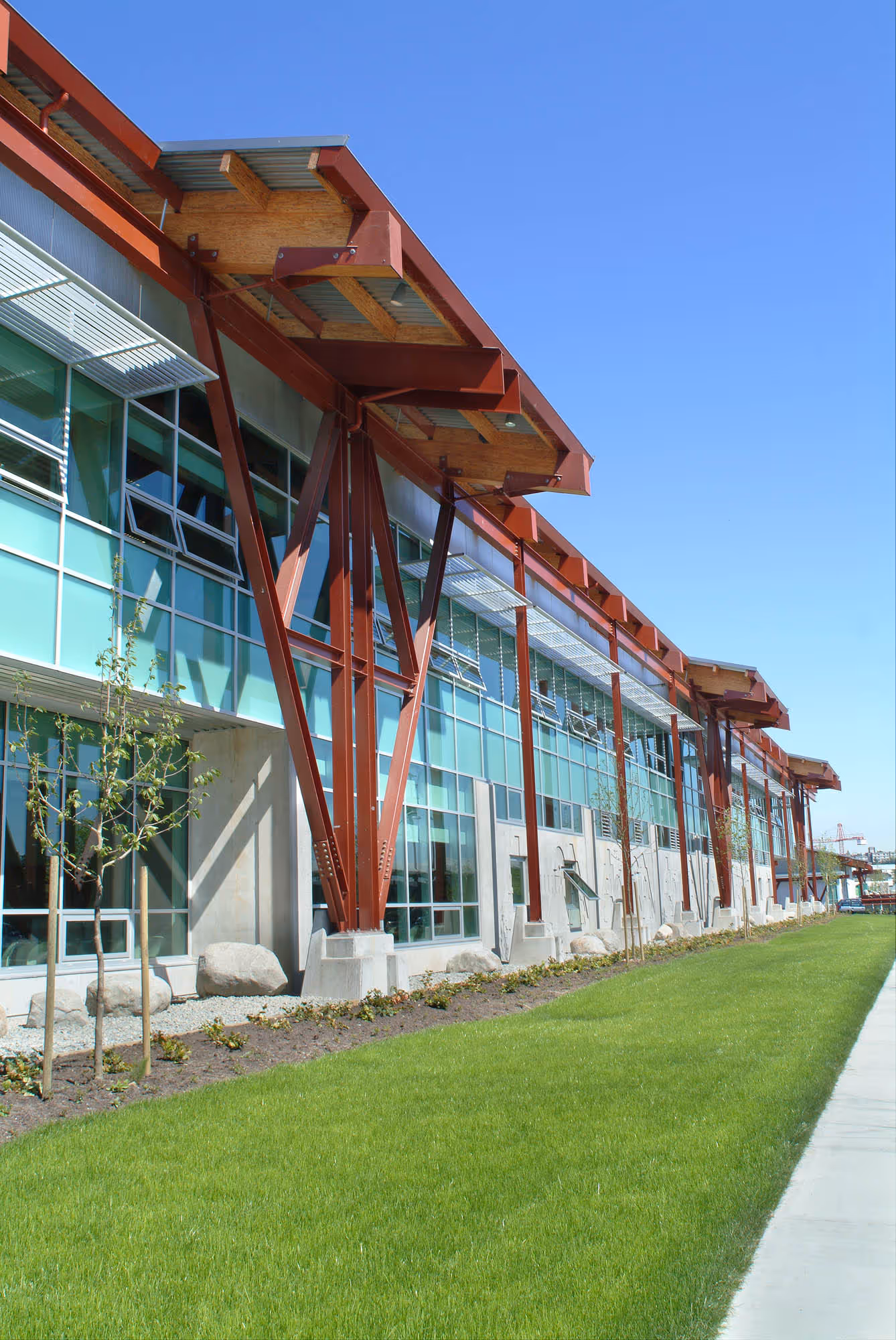 Modern building with large glass windows, red steel beams, small trees, and green lawn under a clear blue sky.