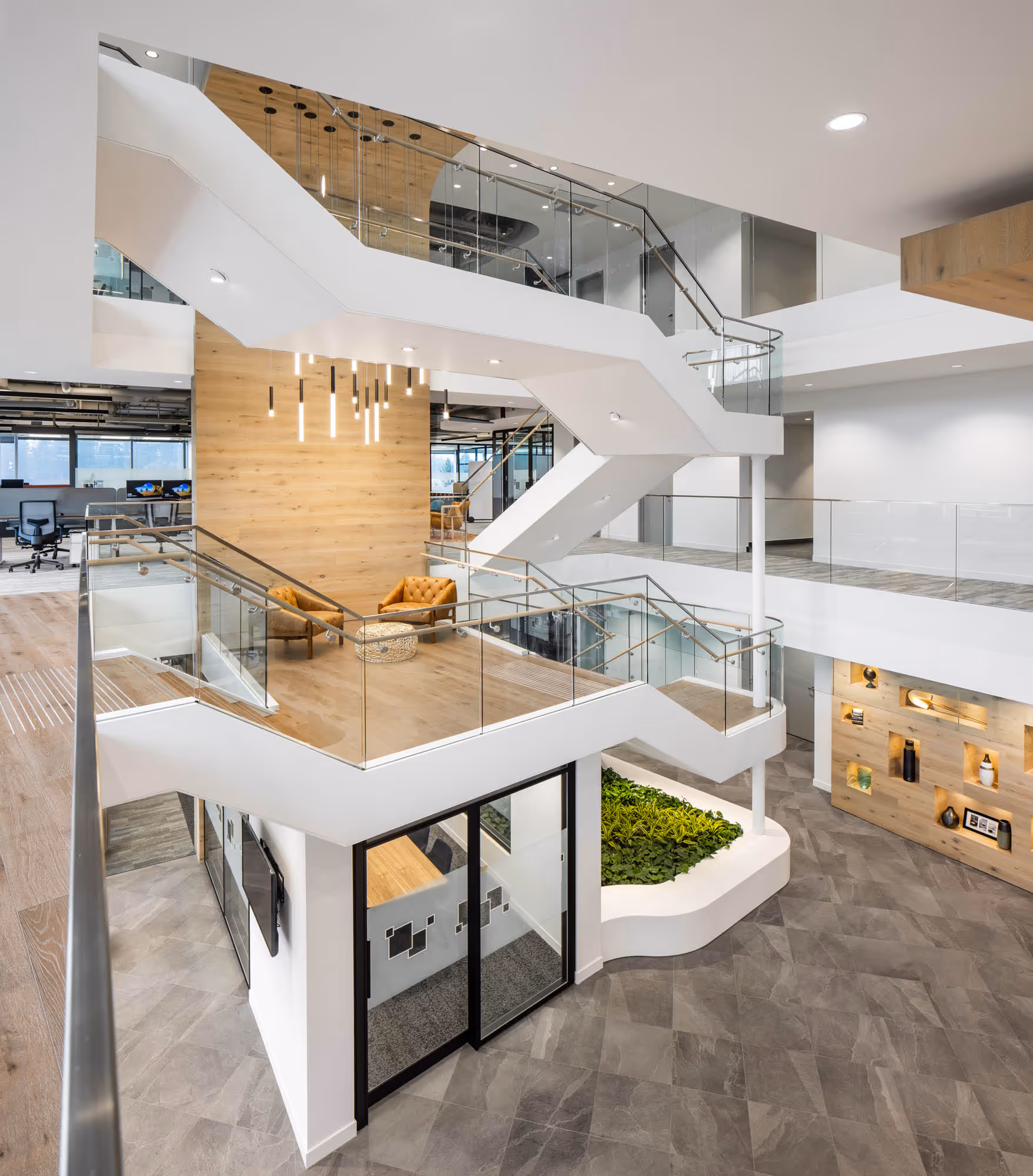 Modern office interior with white staircases, glass railings, wood accents, seating area with brown chairs, and a green plant feature.
