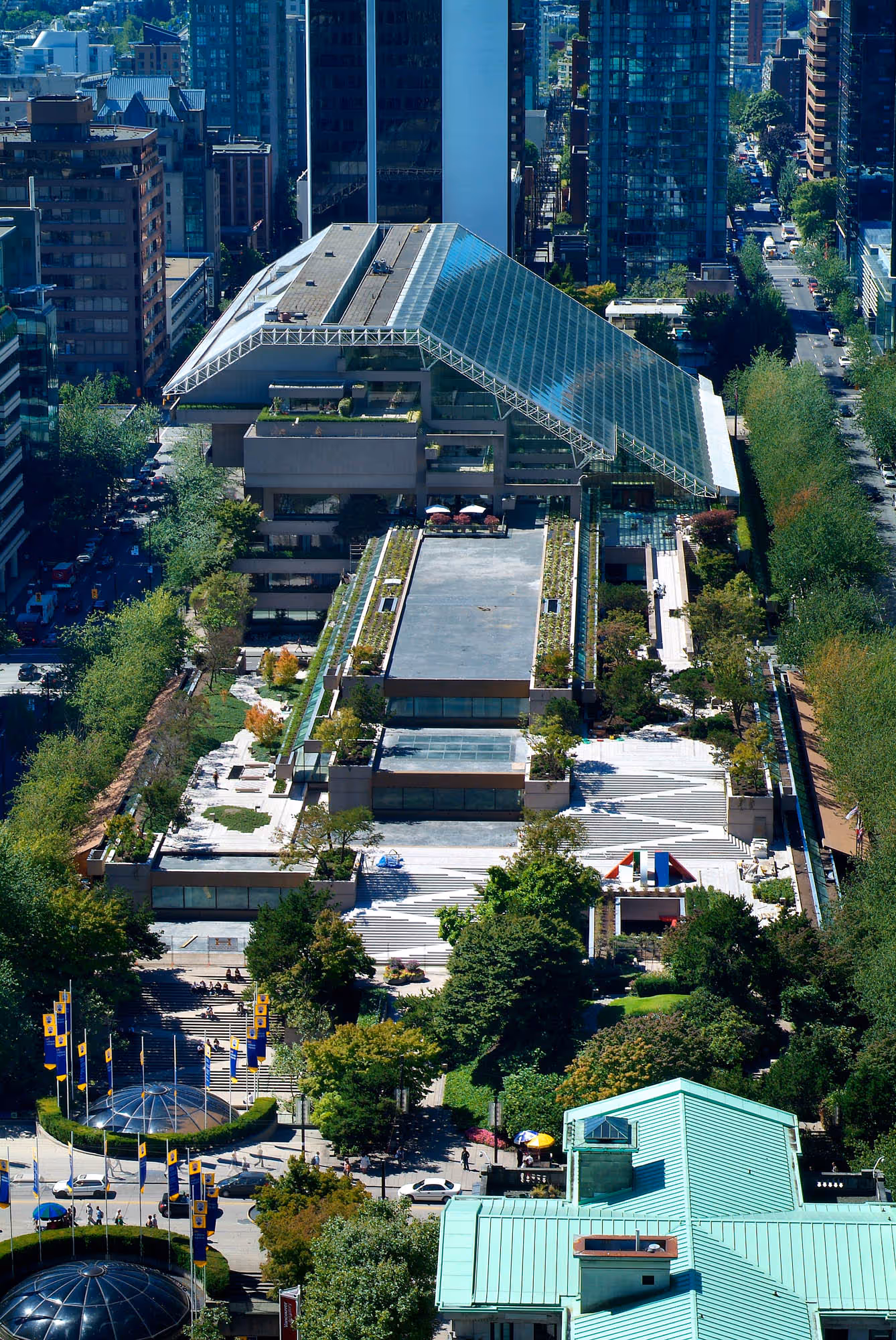Aerial view of Robson Square in Vancouver featuring a modern building with a large glass roof, surrounded by greenery, trees, and urban high-rise buildings.