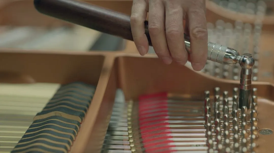 A hand uses a tuning lever to adjust the pins inside a grand piano. The interior strings and hammers are visible, evoking the meticulous craftsmanship akin to Certified Steinway Restorations.