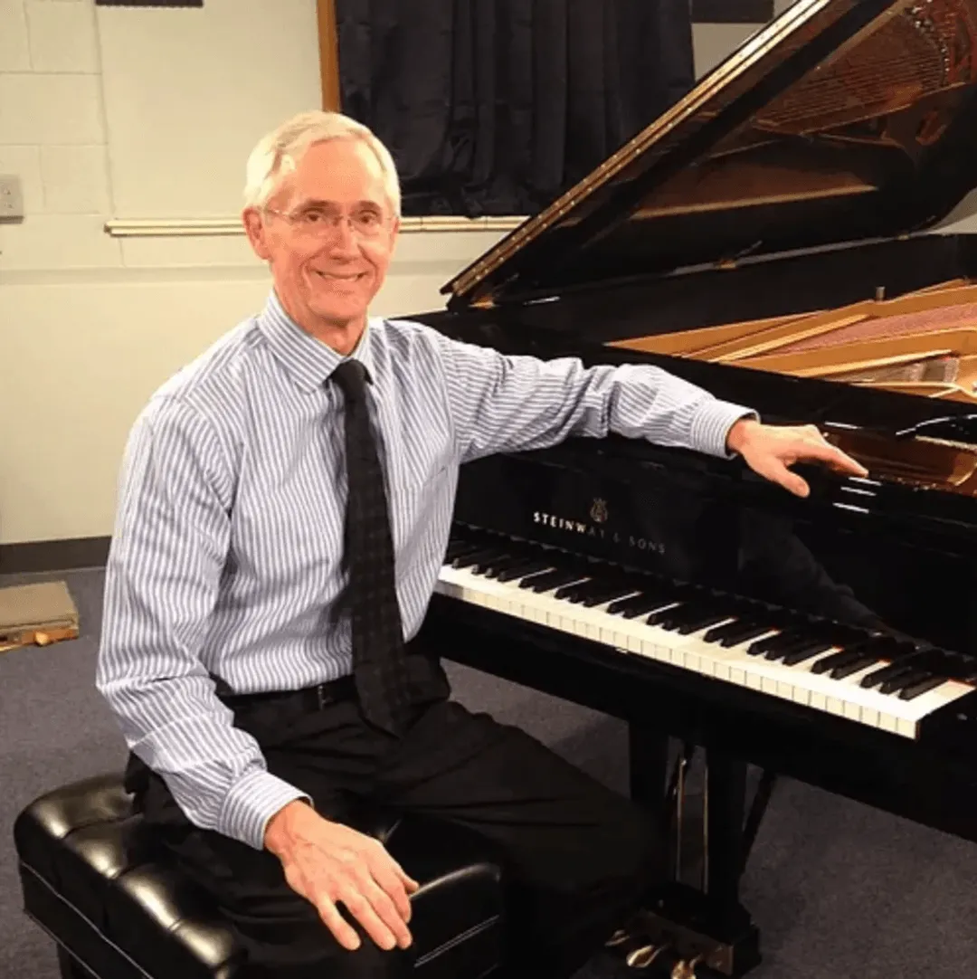 An elderly man in a striped shirt and tie sits on a piano bench, smiling, with one hand on the elegant piano, skillfully restored through Certified Steinway Restorations.