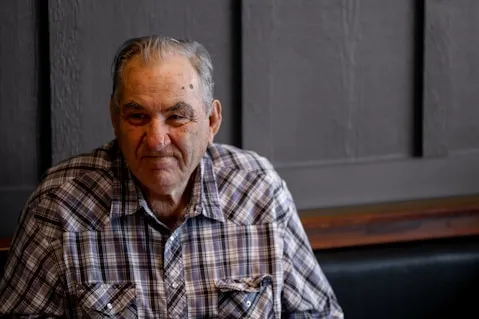 An elderly man with gray hair and a plaid shirt sits indoors against a dark, paneled wall.