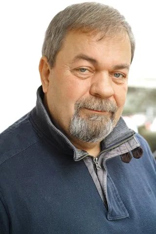 A man with a beard and short hair, clad in a dark zippered sweater, stands confidently against a white background.