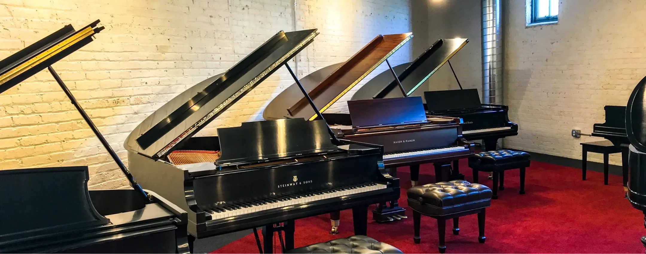 A row of grand pianos with open lids graces the red carpeted floor, set against a white brick wall, showcasing Certified Steinway Restorations crafted with genuine Steinway parts.