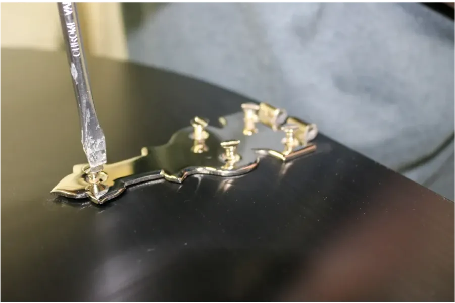A person uses a screwdriver to make precise adjustments, tuning the screws on a gold guitar tailpiece attached to a black guitar body.