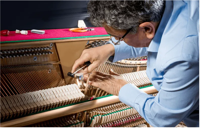 A person wearing glasses and a blue shirt tunes the strings of an upright piano, using a tuning tool on the hammers.