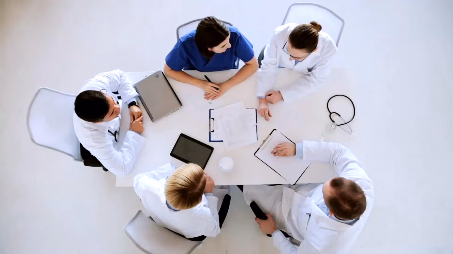 Overhead view of five healthcare professionals in lab coats and scrubs sitting around a table reviewing documents and digital devices.