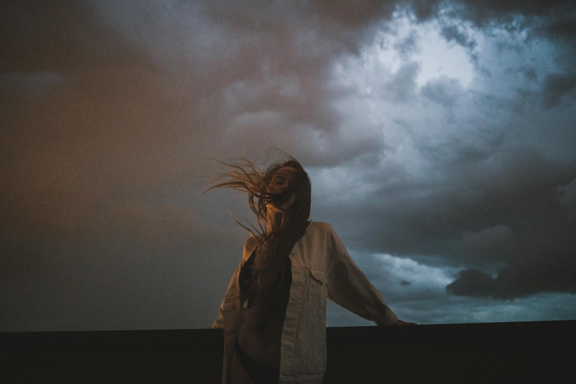 Person with long hair blowing in the wind standing against a dark, cloudy sky at dusk.