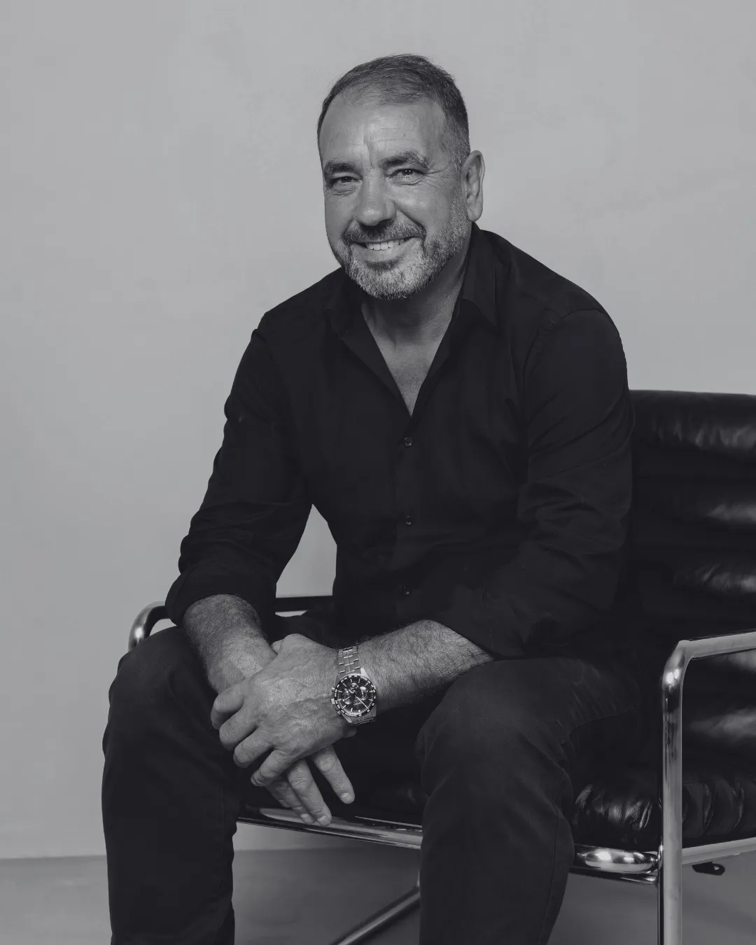 Homem sorridente sentado em cadeira de escritório com camisa preta e relógio de pulso.