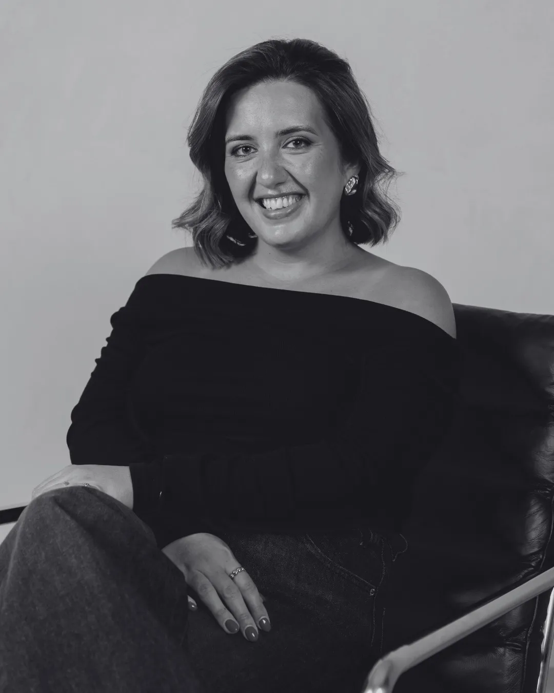 Mulher sorridente com cabelo ondulado, usando blusa preta de ombros à mostra, sentada em cadeira de couro.