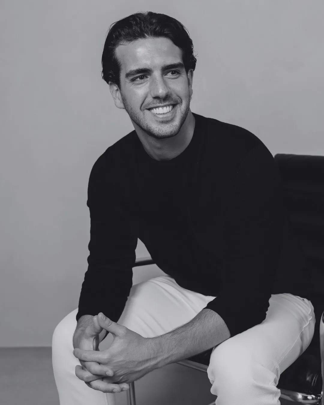 Homem sorridente sentado, vestindo suéter preto e calça branca.