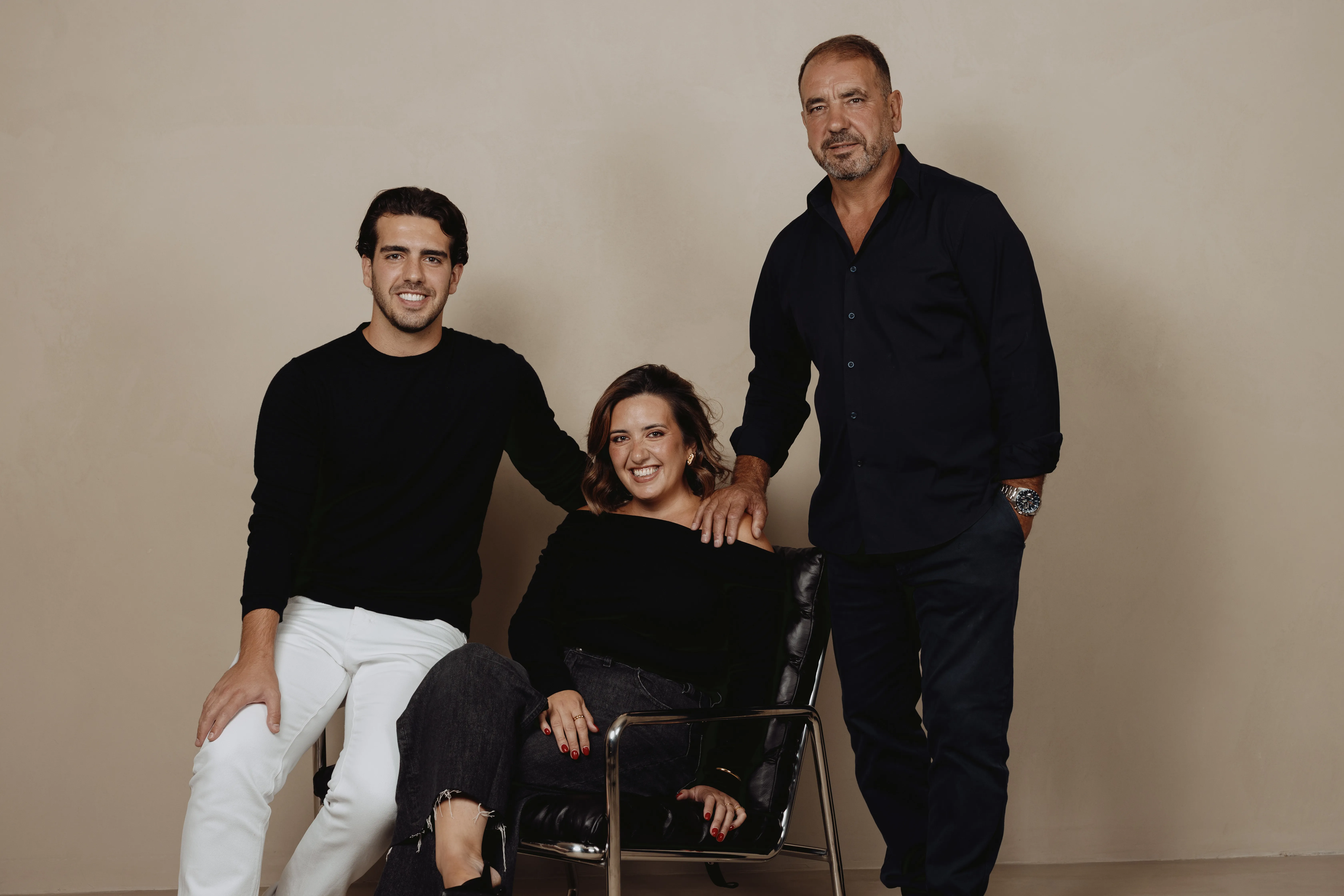 Três pessoas, dois homens e uma mulher sorrindo em estúdio com fundo bege; mulher está sentada em cadeira preta, homens estão ao lado dela.