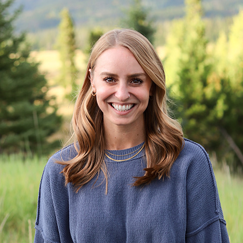 Person with long wavy blonde hair, wearing a blue sweater and layered necklace, smiling outdoors with pine trees and mountains in the background.