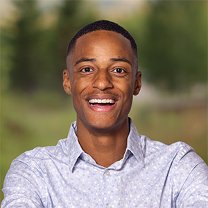 Person with short hair wearing a light blue patterned shirt, smiling widely outdoors with blurred greenery and trees in the background.