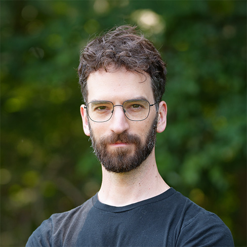 Person with curly dark hair and full beard wearing round wire‑frame glasses and a black T‑shirt, standing outdoors with green foliage behind.