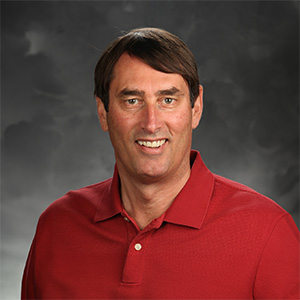Person with short dark brown hair, wearing a red polo shirt, smiling in front of a dark studio background with soft cloud patterns.