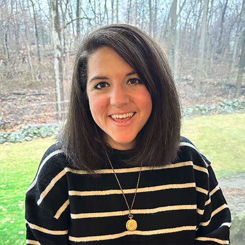 Person with shoulder‑length dark brown hair wearing a black and white striped sweater and a gold pendant necklace, smiling near a window with a view of trees and grass outside.