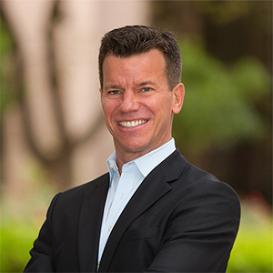Person with short dark hair wearing a black suit jacket and white dress shirt, standing with arms crossed and smiling outdoors with blurred greenery and architecture behind.