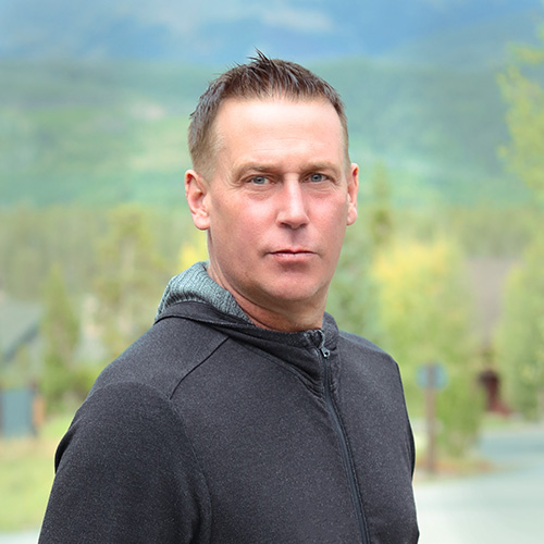 Person with short hair wearing a dark hooded jacket, posing outdoors with a backdrop of mountains, trees and a road.
