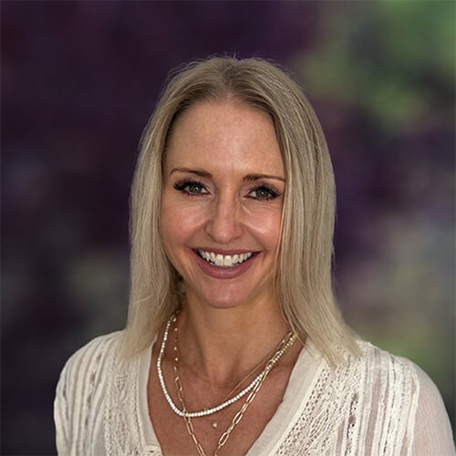 Person with straight shoulder‑length blonde hair wearing a cream textured top and layered necklaces, smiling against a blurred purple and green backdrop.