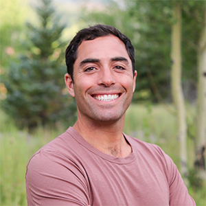 Person with short dark hair wearing a mauve T‑shirt, standing with arms crossed and smiling outdoors among trees and greenery.