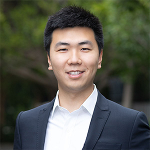 Person with short black hair wearing a dark suit jacket over a white shirt, smiling outdoors with blurred trees in the background.