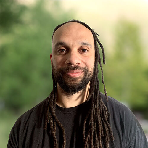 Person with a shaved head and long dreadlocks wearing a black shirt and beard, posing outdoors with a blurred green backdrop.