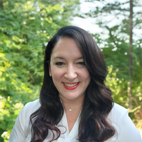 Person with long wavy dark hair wearing a white blouse and red lipstick, smiling outdoors with lush green foliage behind.