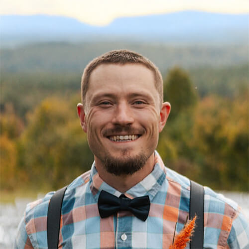 Person with very short hair and goatee wearing a colorful plaid shirt, dark bow tie and suspenders, smiling outdoors with a scenic mountain and forest backdrop.