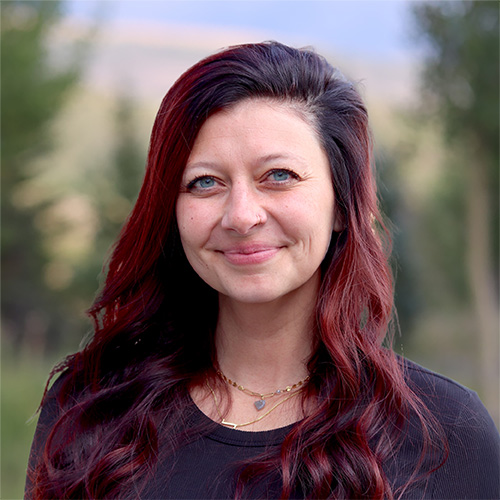 Person with long wavy hair with dark roots and burgundy ends wearing a black top and layered gold necklaces, smiling outdoors with blurred natural scenery behind.