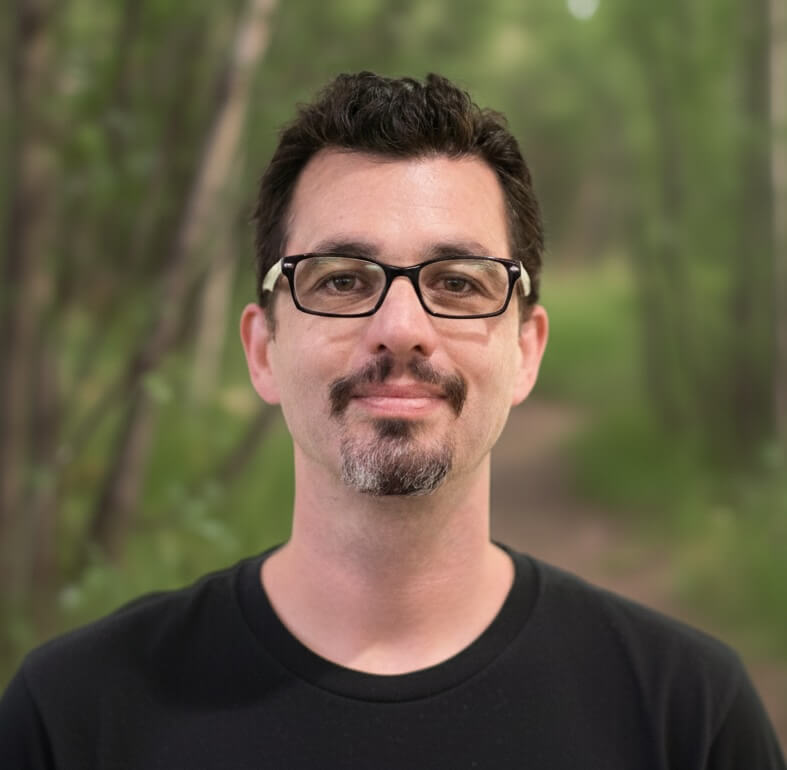 A man with short dark hair, glasses, and a goatee smiles slightly, wearing a black shirt in front of a nature background.