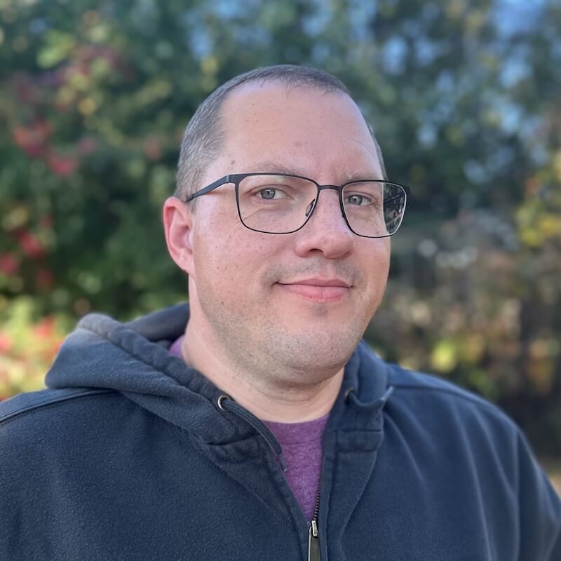 Person with short hair and rectangular glasses wearing a plaid shirt over a dark T‑shirt, smiling broadly with a blurred outdoor background of trees and hills.