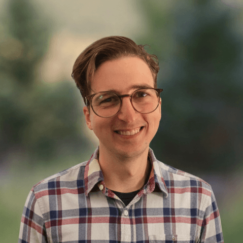 Portrait of a smiling person with short hair and glasses, wearing a plaid button-down shirt against a blurred green background.