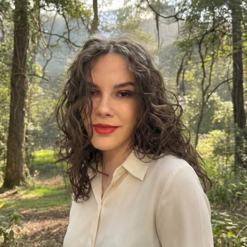 Person with curly dark hair wearing a light-colored blouse, standing outdoors in a wooded setting.