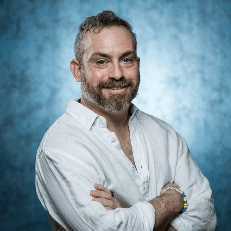 Professional headshot of smiling man with short gray hair and beard, wearing light button-down shirt against blue backdrop.