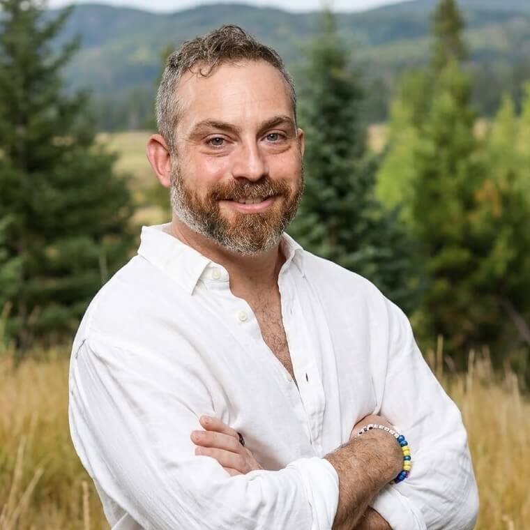 A smiling man with a beard and salt-and-pepper hair stands outdoors with arms crossed, wearing a white button-down shirt.