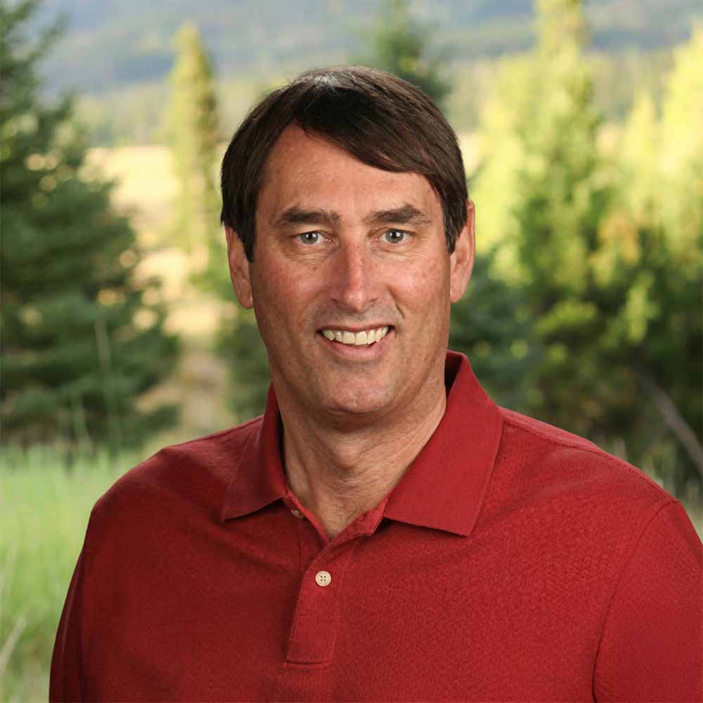 Headshot of a smiling man wearing a red polo shirt, standing outdoors with blurred trees and hills in the background, natural lighting, casual professional portrait with greenery and mountains behind him.