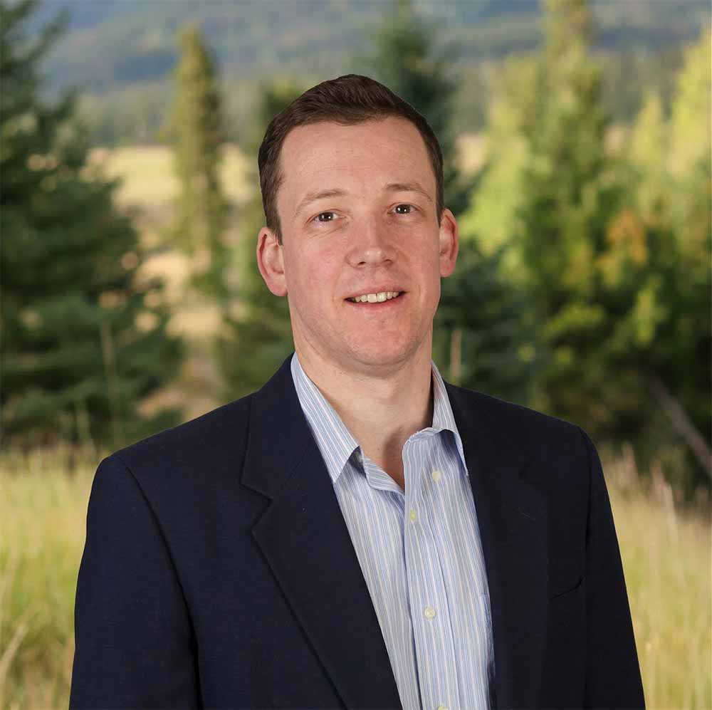 Headshot of a man wearing a navy blazer and light blue striped shirt, standing outdoors with trees and hills softly blurred behind him, professional portrait with natural lighting and scenic background.