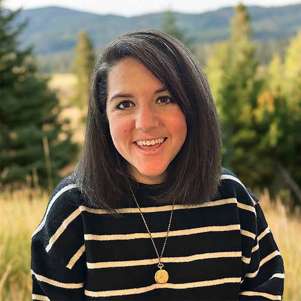 Headshot of a smiling woman with shoulder-length dark hair wearing a black sweater with white stripes and a gold pendant necklace, standing outdoors with blurred trees and mountains in the background.
