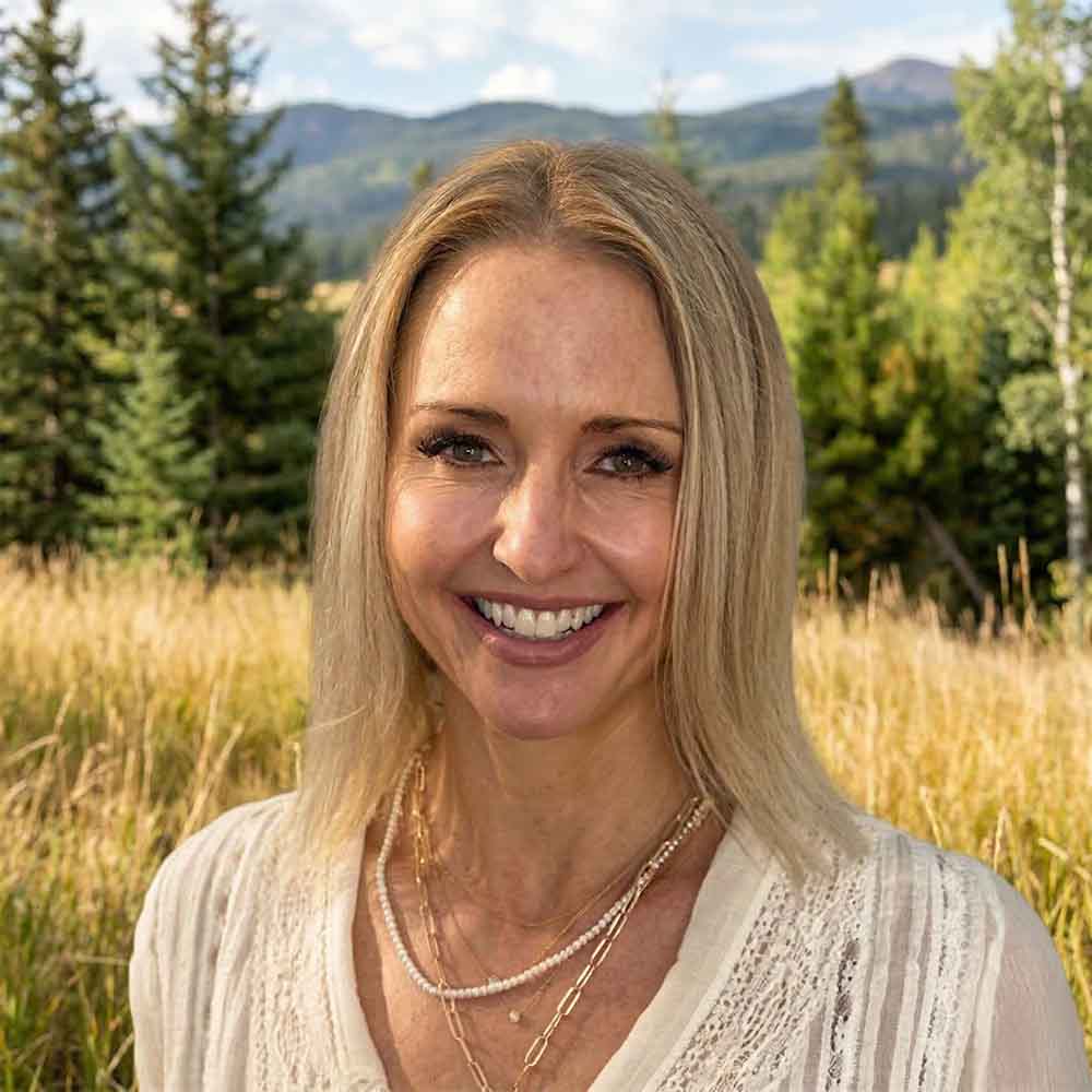 Headshot of a smiling woman with long blonde hair wearing a light-colored blouse and layered necklaces, standing outdoors with tall grass, evergreen trees, and mountains softly blurred in the background.