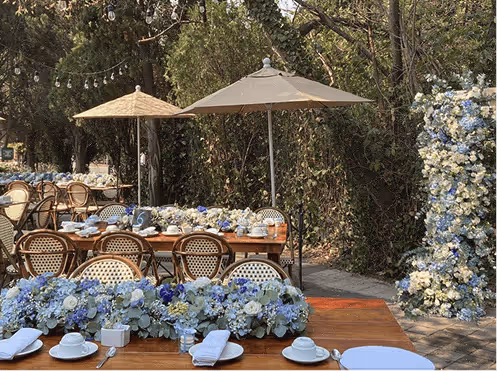 Mesas al aire libre decoradas con arreglos florales blancos y azules, con sombrillas y sillas de mimbre en un jardín.