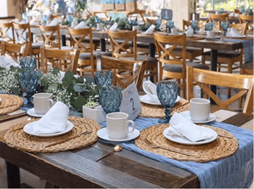 Mesa rústica elegantemente puesta con platos, tazas, copas azules y servilletas blancas, adornada con centros de mesa de arreglos florales verdes.