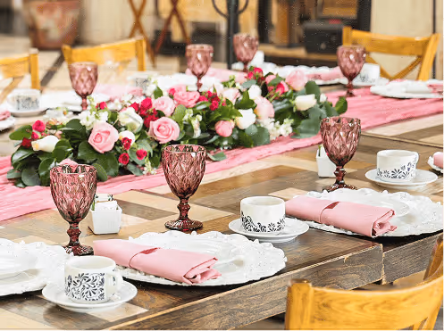 Mesa rústica decorada con copas de vino rosa, tazas blancas con diseño negro, servilletas rosadas y un centro floral de rosas blancas y rosas.