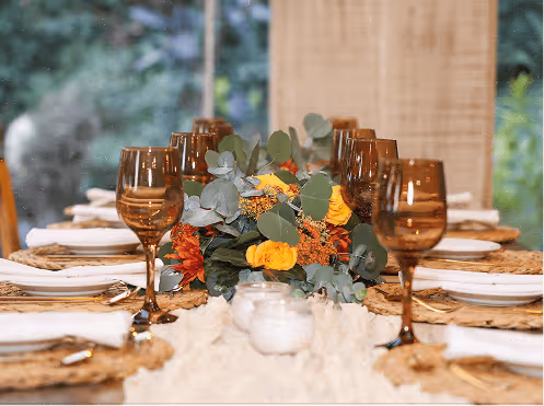 Mesa elegantemente puesta con copas de vino marrón, centros de mesa con flores amarillas y naranjas, y vajilla blanca.
