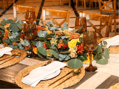 Mesa decorada para evento con vajilla dorada, copas marrones y centro de mesa con flores amarillas, rojas y follaje verde.
