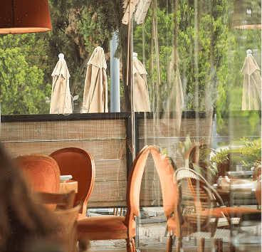 Interior de cafetería con sillas naranjas y mesas visibles a través de un cristal que refleja sombrillas cerradas y vegetación exterior.